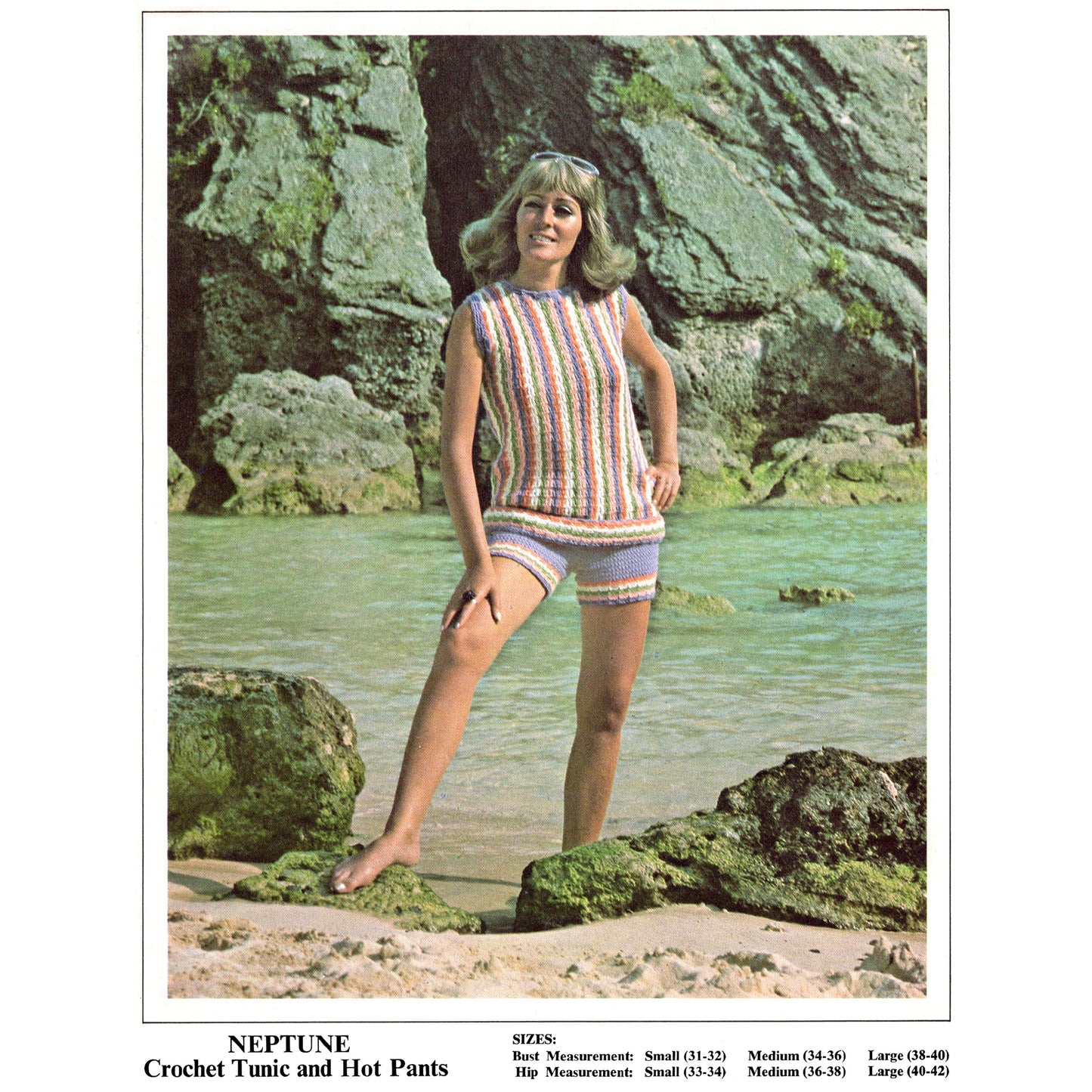 Woman in a crochet tunic and hot pants standing on a rocky beach with water in the background.
