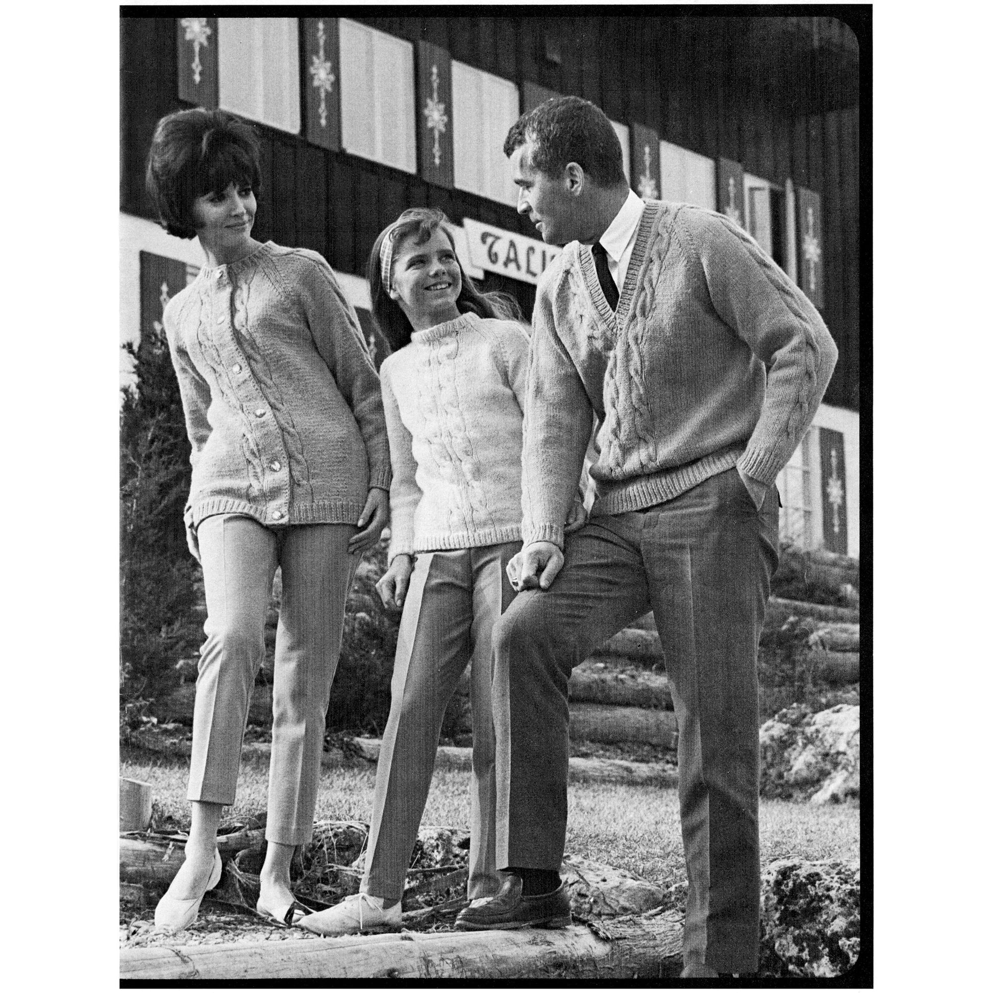 Three people wearing knitted sweaters standing outdoors with a wooden building in the background.