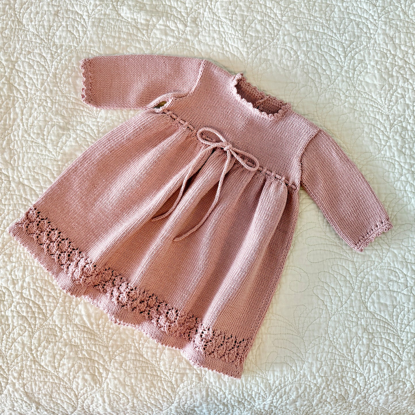 A dusty pink hand-knit baby dress with long sleeves, a scalloped neckline, a drawstring bow at the waist, and a floral lace hem, laid flat on a textured cream quilted blanket.