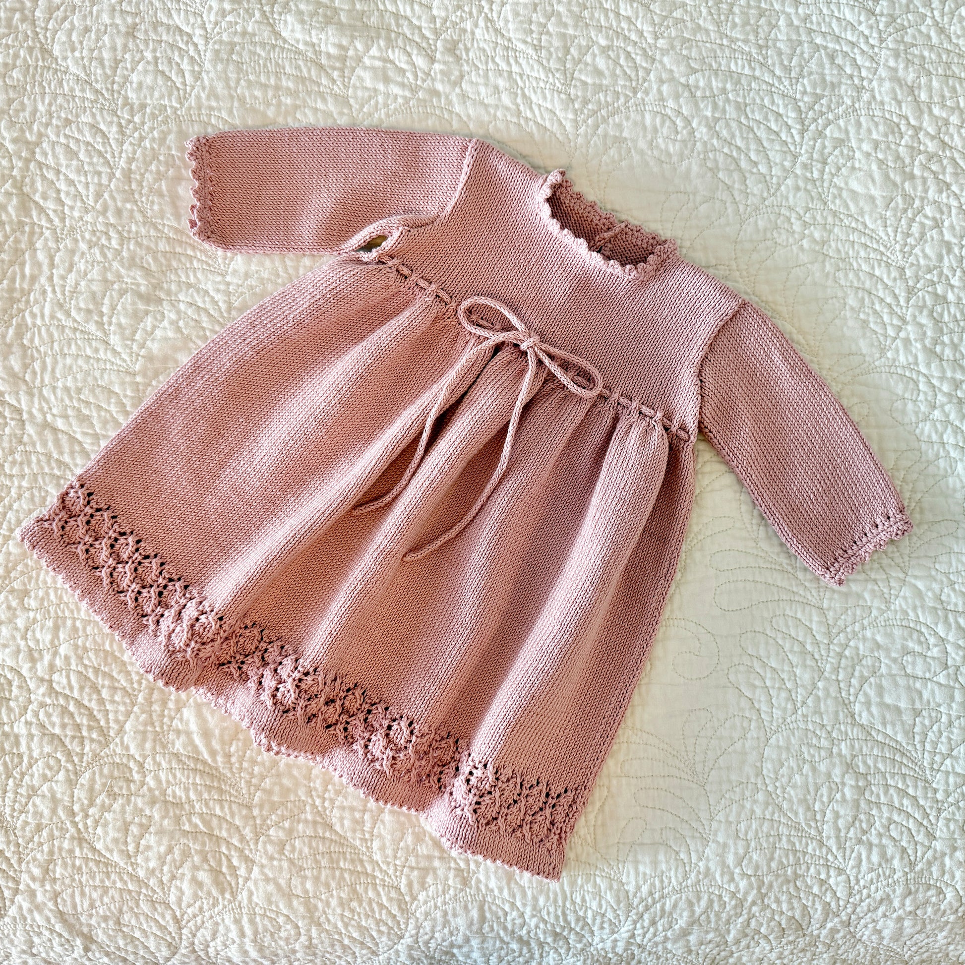 A dusty pink hand-knit baby dress with long sleeves, a scalloped neckline, a drawstring bow at the waist, and a floral lace hem, laid flat on a textured cream quilted blanket.
