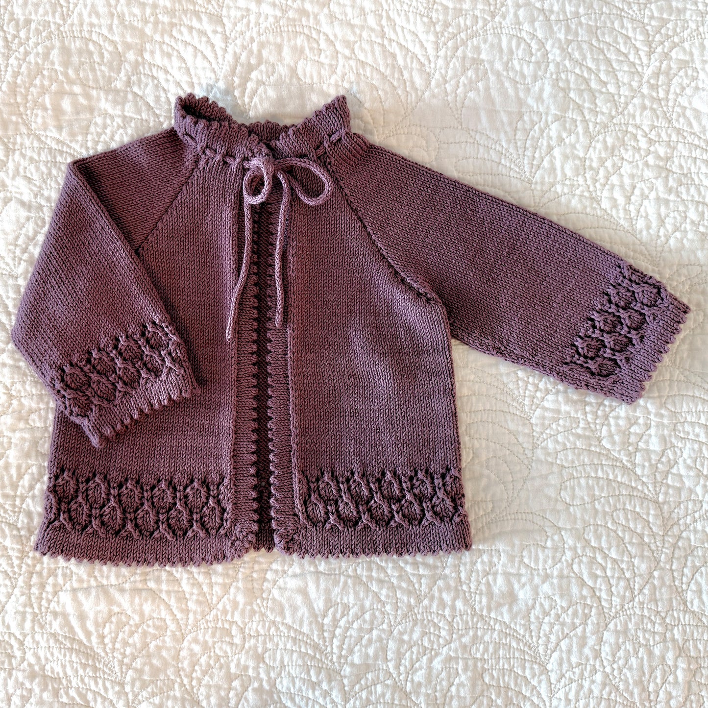 A handmade, long-sleeve knit cardigan in a dusty plum purple color, laid flat on a white quilted blanket. The cardigan features a ruffled collar with a tie closure, and decorative lace-like patterns on the cuffs and hem.