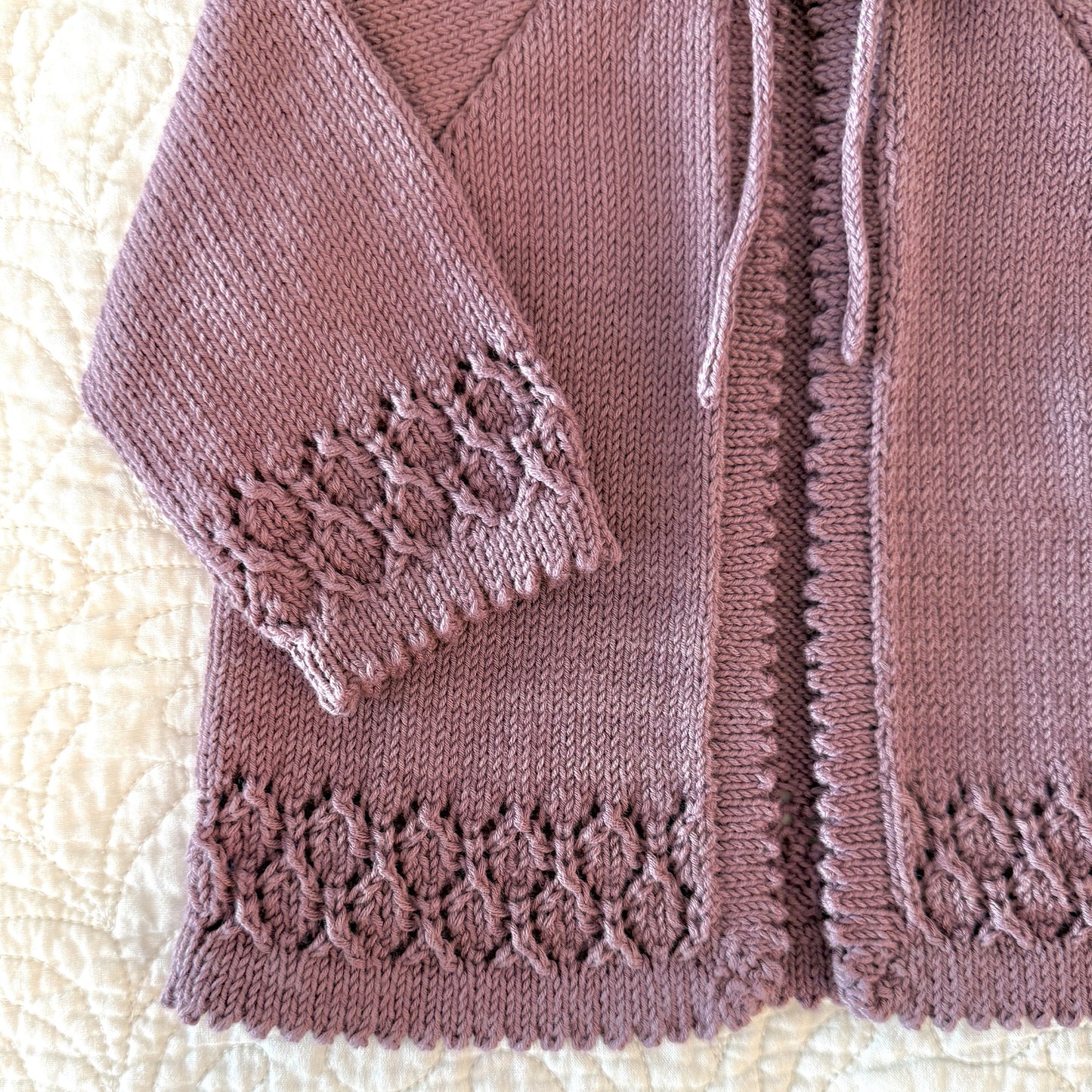 A close-up of a knitted cardigan in a dusty mauve or muted rose color, laid flat on a cream quilted blanket. The cardigan features a textured cable-knit pattern, a drawstring closure, and decorative lace-like knit trim at the cuffs and hem.
