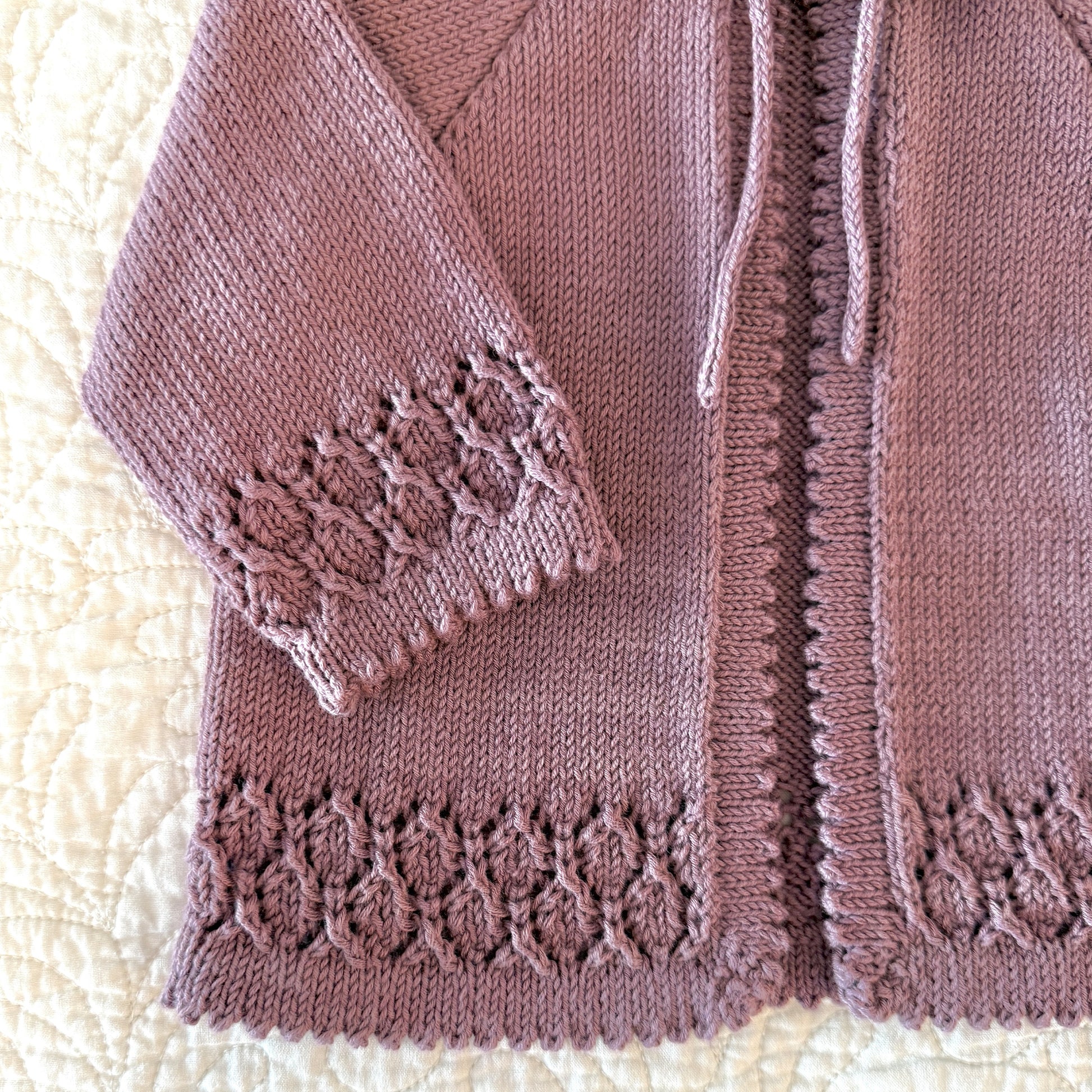 A close-up of a knitted cardigan in a dusty mauve or muted rose color, laid flat on a cream quilted blanket. The cardigan features a textured cable-knit pattern, a drawstring closure, and decorative lace-like knit trim at the cuffs and hem.