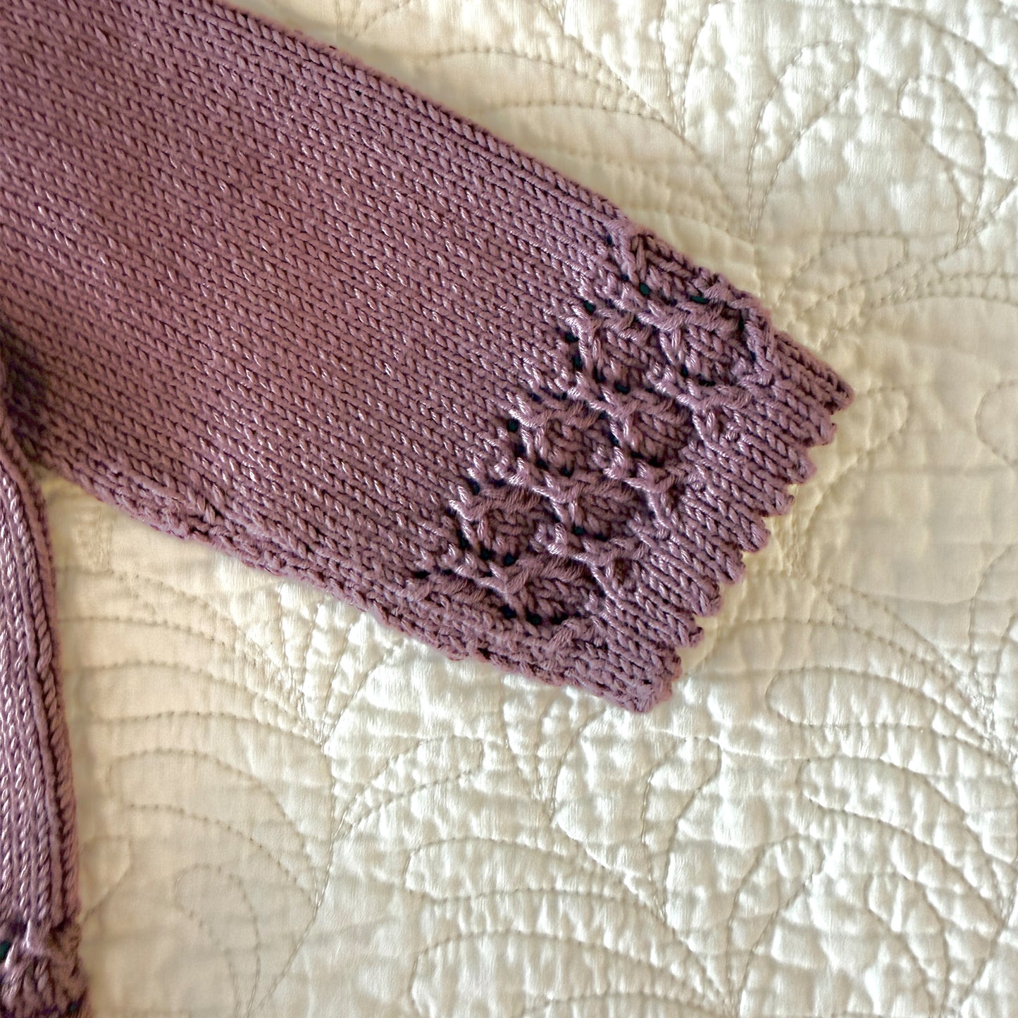 A close-up of a knitted garment sleeve in a dusty mauve-purple color, featuring a textured floral lace cuff, laid flat on a cream-colored quilted blanket.