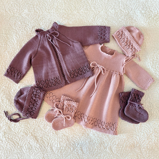 A handmade baby clothing set laid flat on a cream textured blanket, featuring a dusty mauve knit cardigan, a light pink dress, matching bonnets, booties, and mittens.