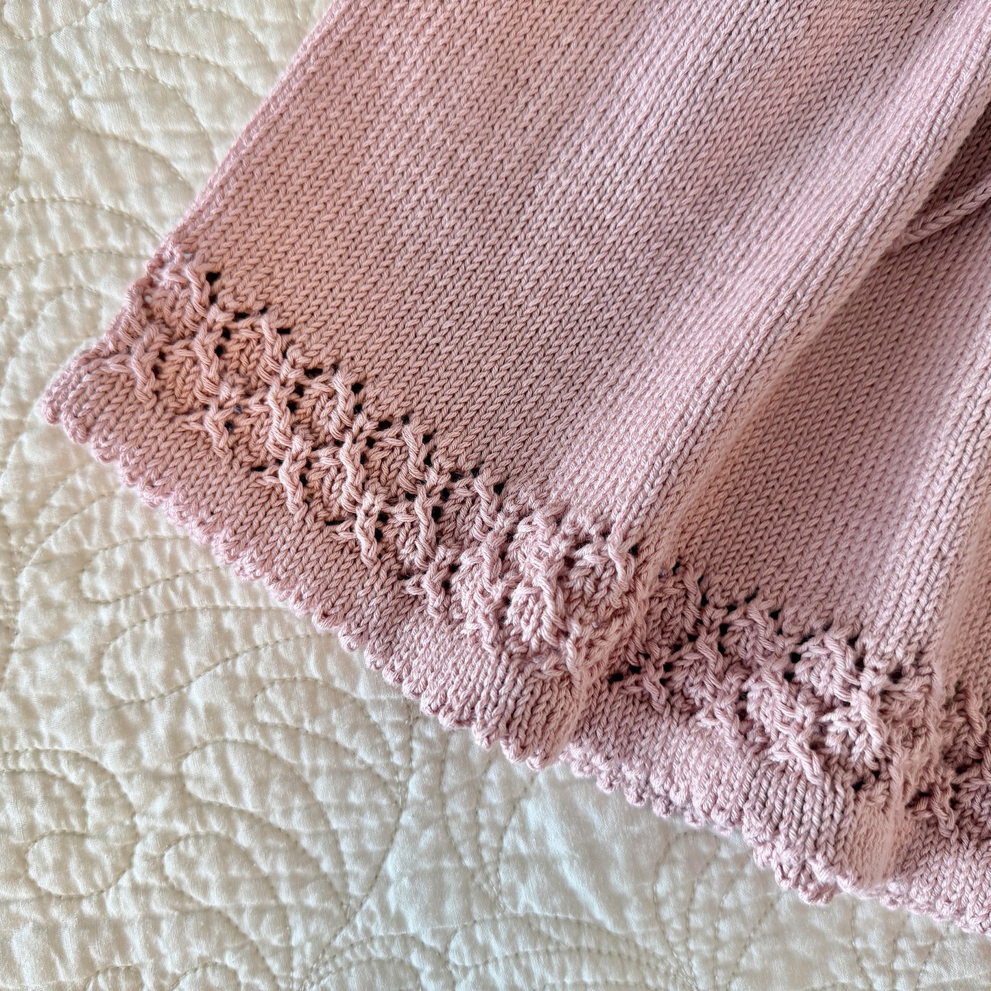 A close-up of a dusty pink knitted textile, featuring a textured floral lace trim along one edge, laid flat on a cream-colored quilted surface.