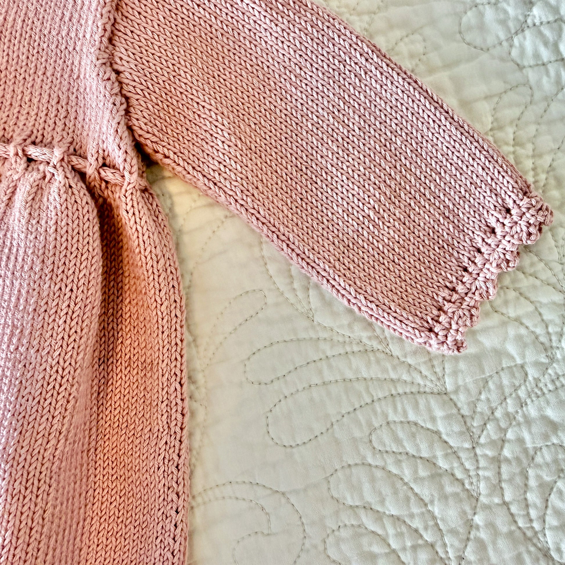 Close-up of a pink knitted garment laid on a cream quilted blanket, showing the knit texture and sleeve cuff detail.