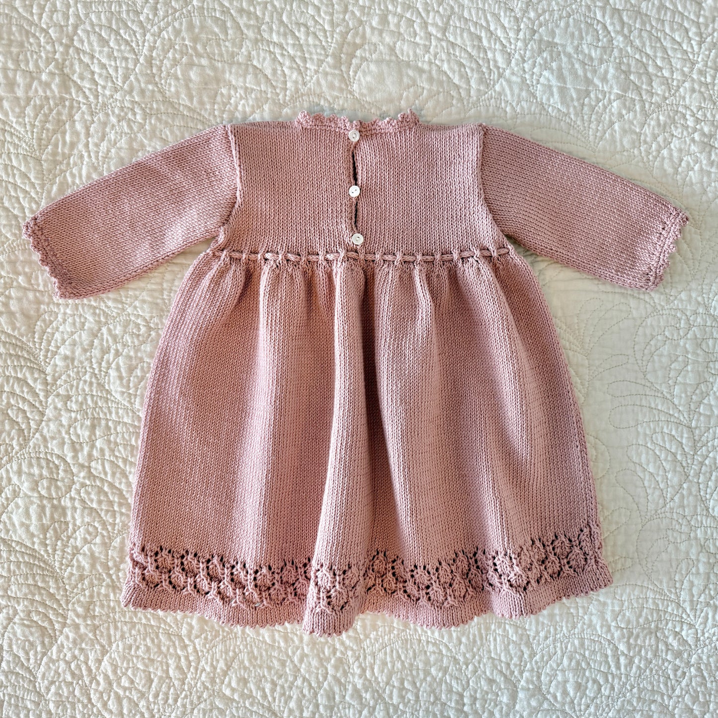 Back view of a dusty pink hand-knitted baby dress with long sleeves, a buttoned neckline, and a decorative lace hem, laid flat on a white quilted blanket.