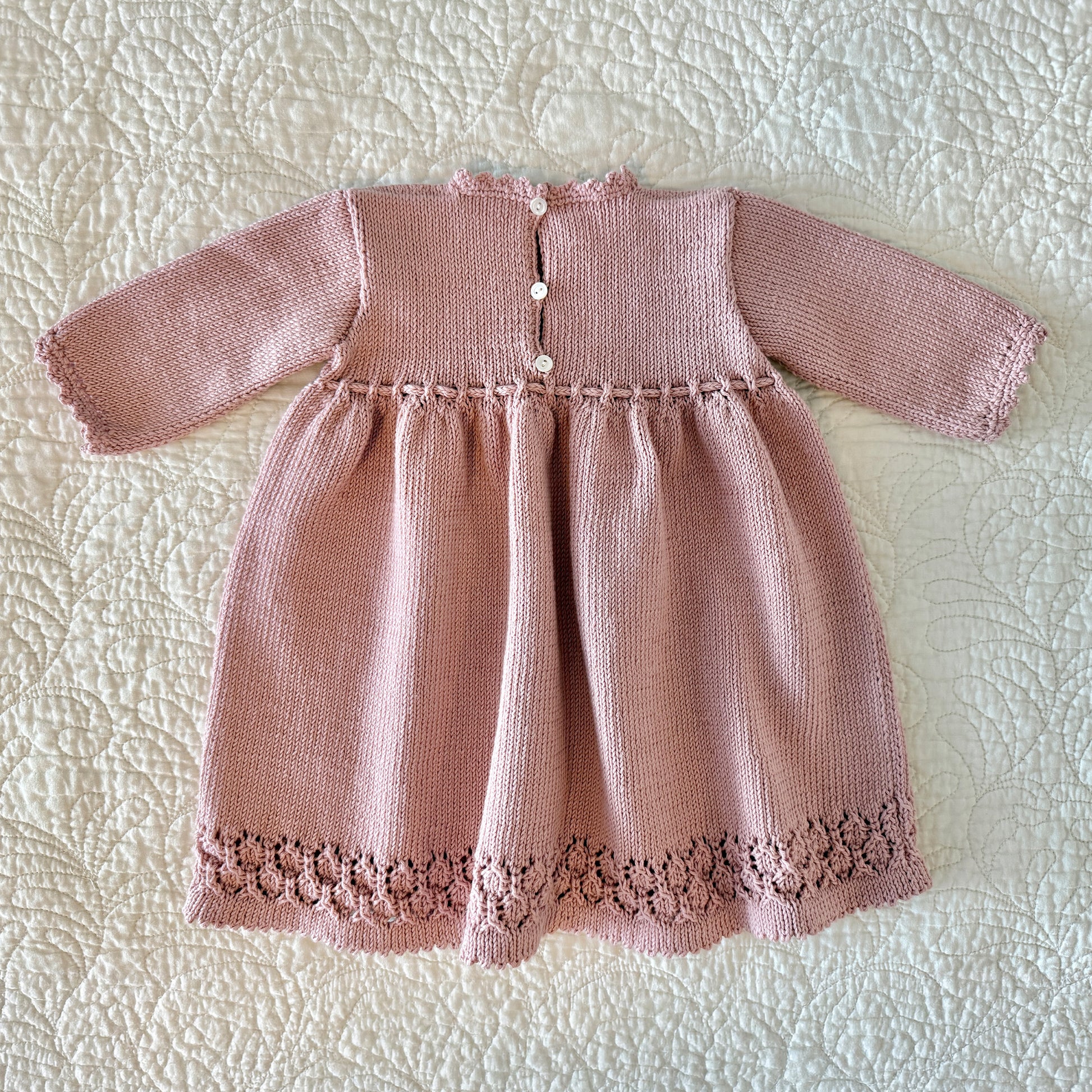 Back view of a dusty pink hand-knitted baby dress with long sleeves, a buttoned neckline, and a decorative lace hem, laid flat on a white quilted blanket.