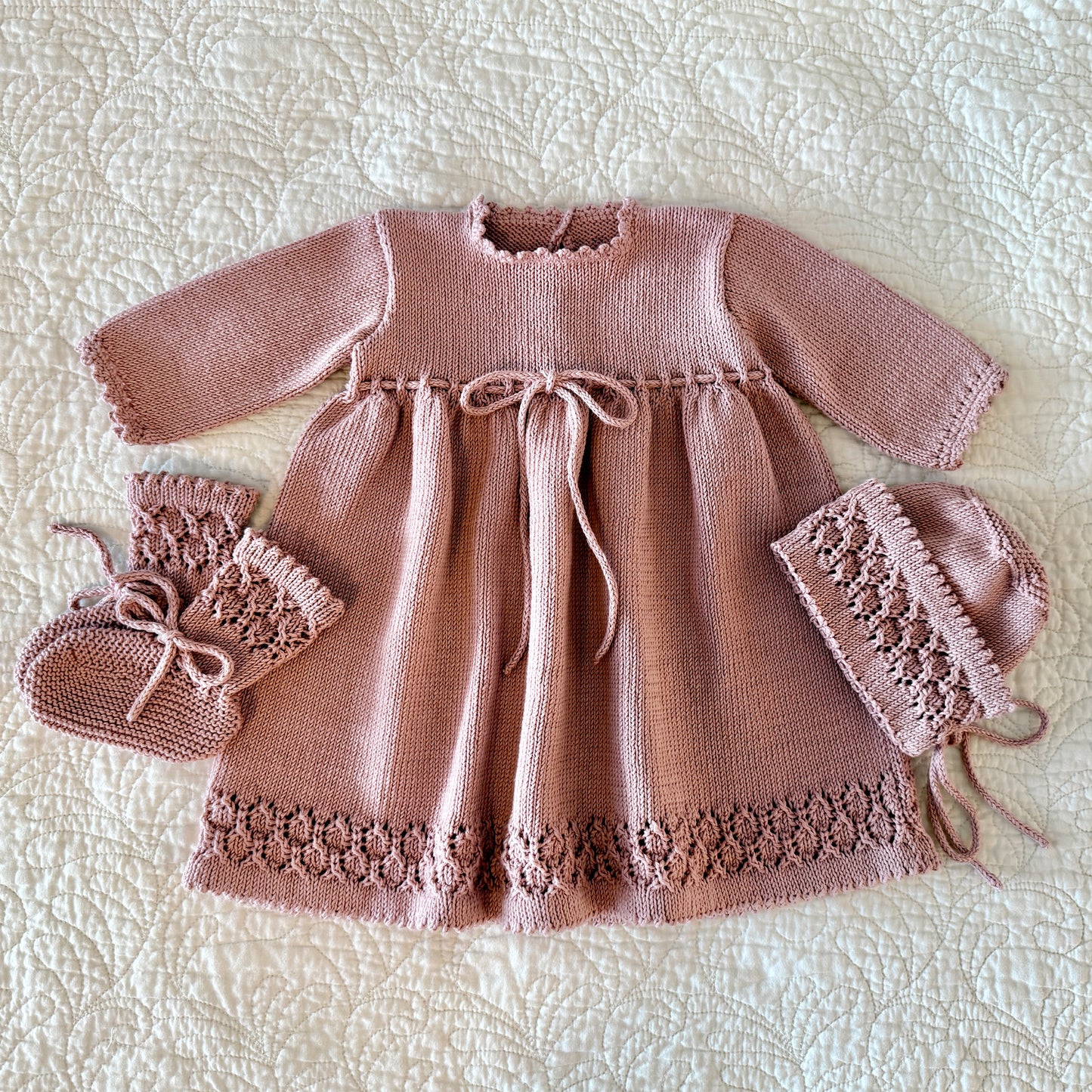 A hand-knitted baby outfit set in dusty rose pink, including a long-sleeve dress with a tie waist, matching booties, and a bonnet, laid flat on a textured cream quilted blanket.