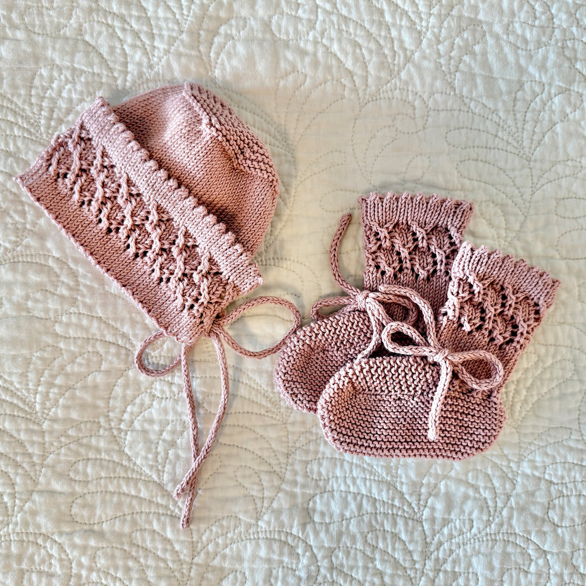 A matching pink knitted baby hat and bootie set laid flat on a white quilted blanket. The hat has lace-like knit detailing and drawstrings, while the booties are knitted with lace panels and bow ties.
