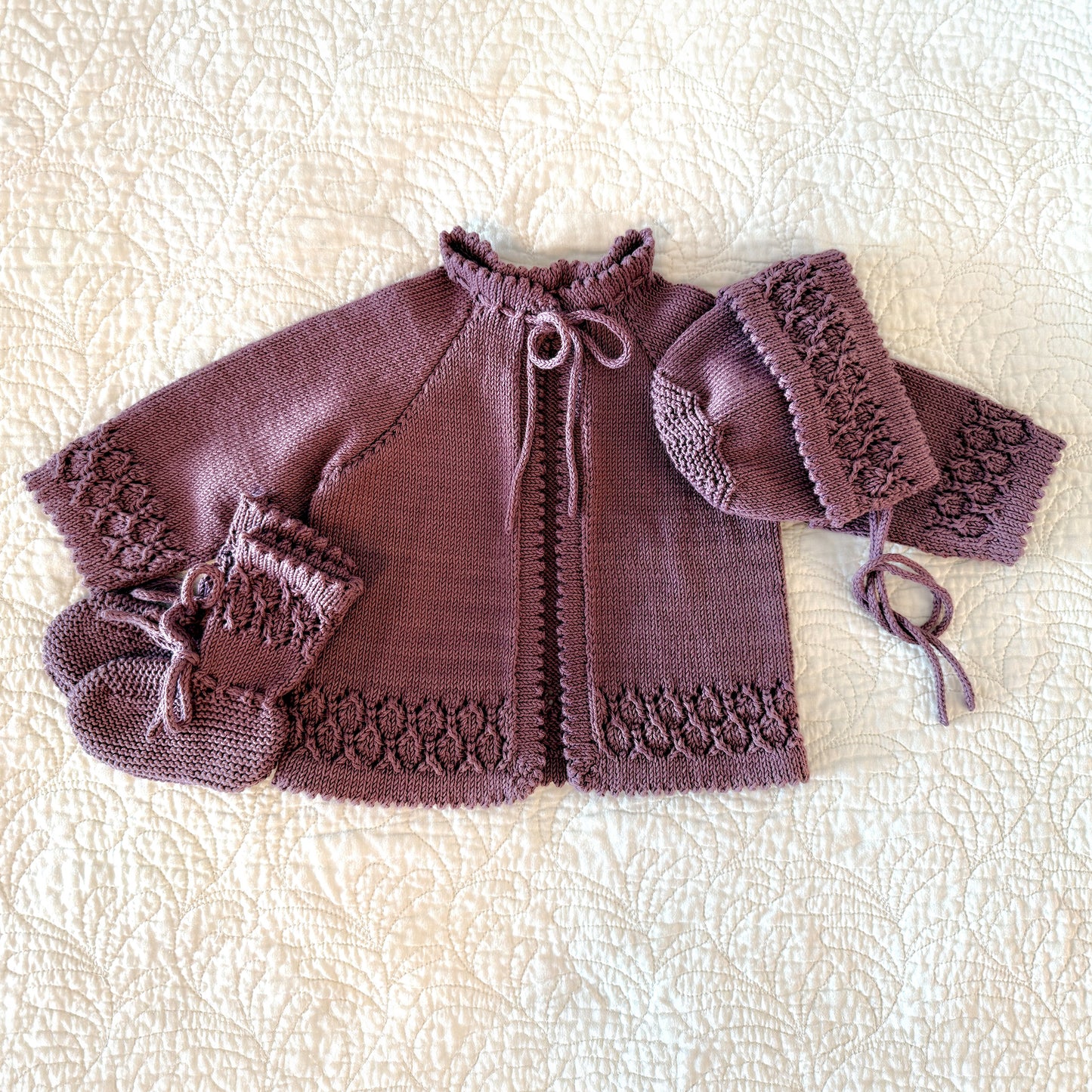 A handmade knit baby outfit set in a dusty mauve-purple color, featuring a short-sleeve cardigan with lace trim, a bonnet, and booties, laid flat on a textured cream quilted blanket.