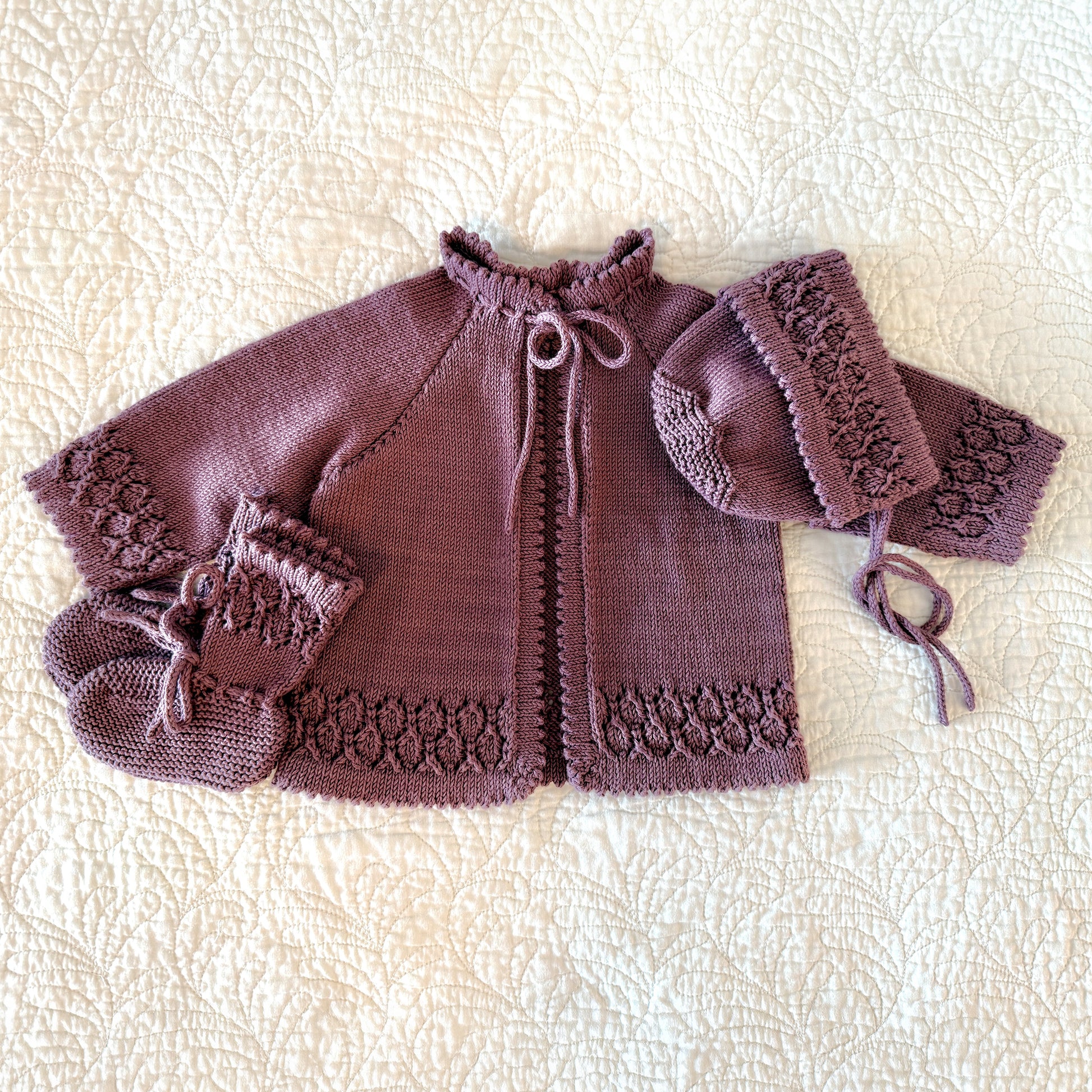 A handmade knit baby outfit set in a dusty mauve-purple color, featuring a short-sleeve cardigan with lace trim, a bonnet, and booties, laid flat on a textured cream quilted blanket.