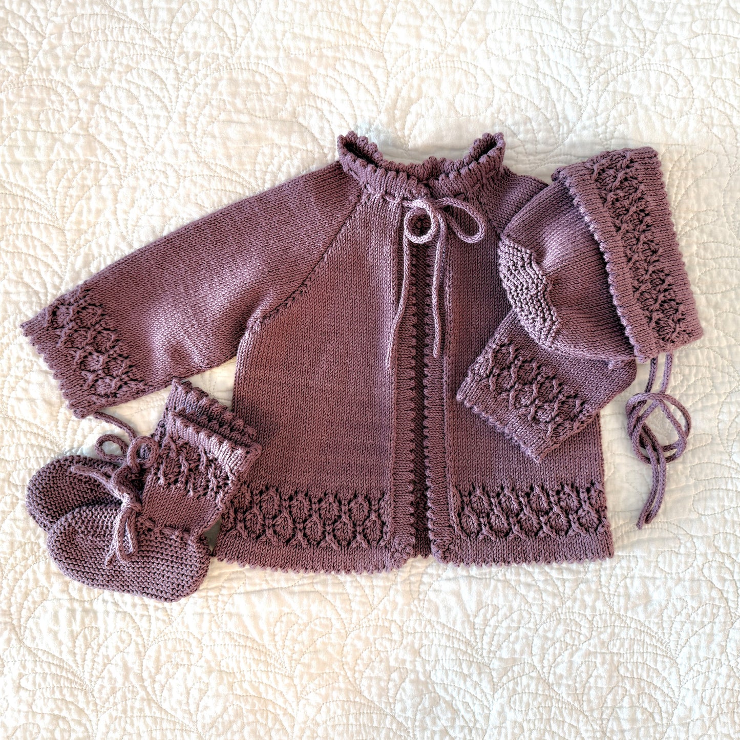 A handmade knit baby outfit set in a dusty mauve-purple color, including a cardigan, bonnet, and booties, laid flat on a cream-colored textured quilted blanket.