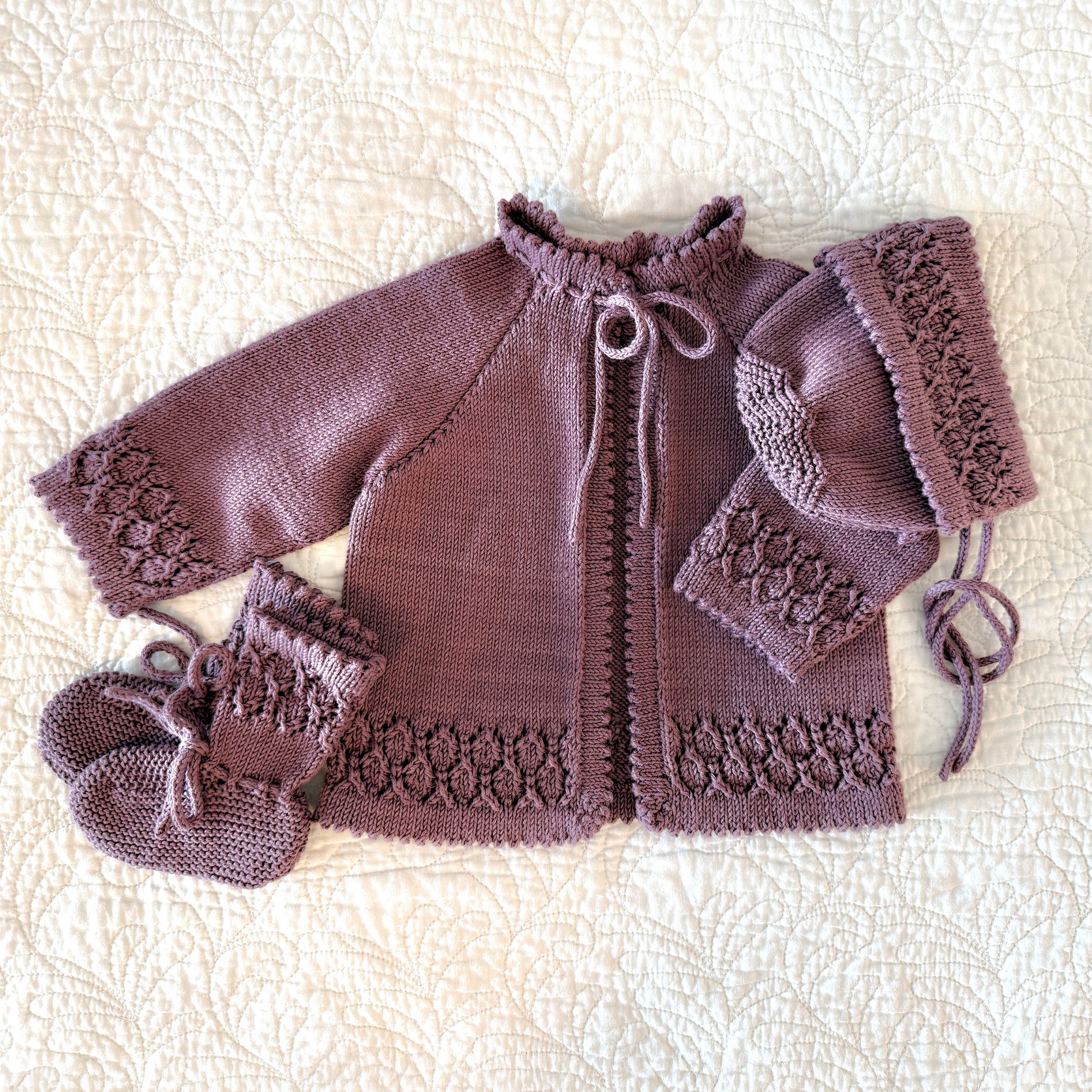 A handmade knit baby outfit set in a dusty mauve-purple color, including a cardigan, bonnet, and booties, laid flat on a cream-colored textured quilted blanket.