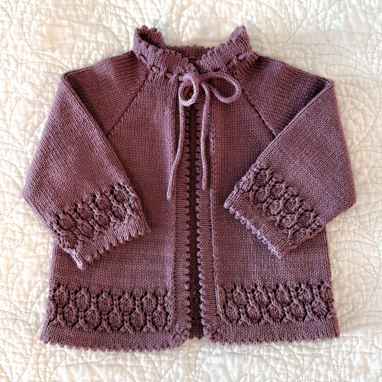 A handmade, hand-knitted baby cardigan in a dusty mauve-purple color, featuring picot edged hems, a drawstring tie, and textured lace patterns on the cuffs and hem, laid flat on a cream quilted blanket.