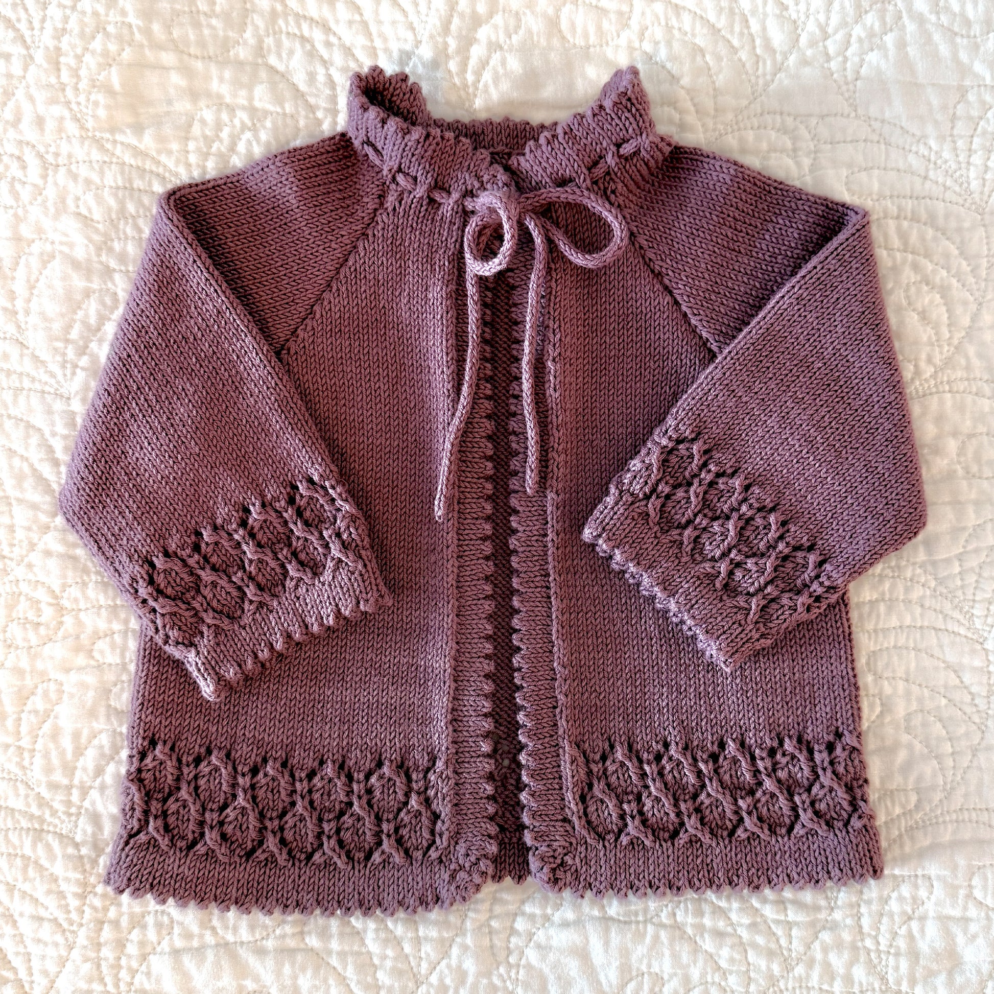 A handmade, hand-knitted baby cardigan in a dusty mauve-purple color, featuring picot edged hems, a drawstring tie, and textured lace patterns on the cuffs and hem, laid flat on a cream quilted blanket.